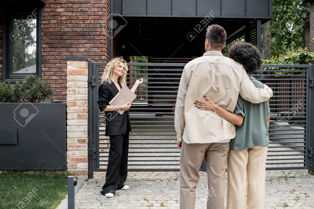 211032240-smiling-blonde-real-estate-agent-with-folder-showing-new-city-cottage-to-multiethnic-couple